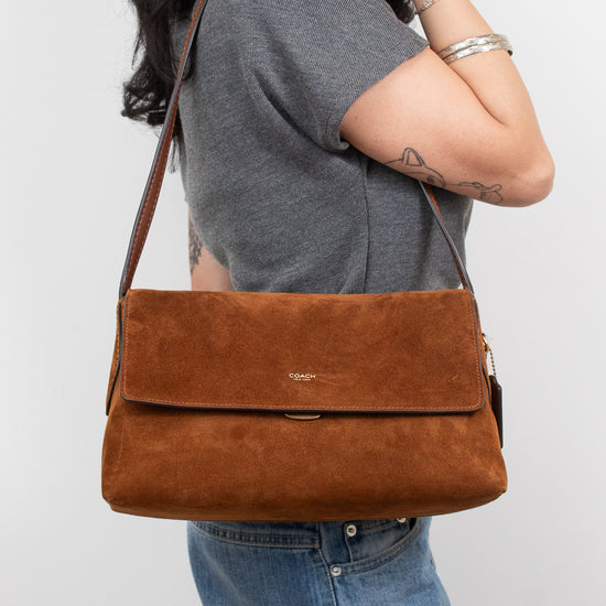 A woman wearing a gray t-shirt and blue jeans carries a brown suede shoulder bag with a flap closure and a small gold logo on the front, set against a plain light gray background.