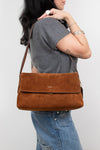 A woman wearing a gray t-shirt and blue jeans carries a brown suede shoulder bag with a flap closure and a small gold logo on the front, set against a plain light gray background.
