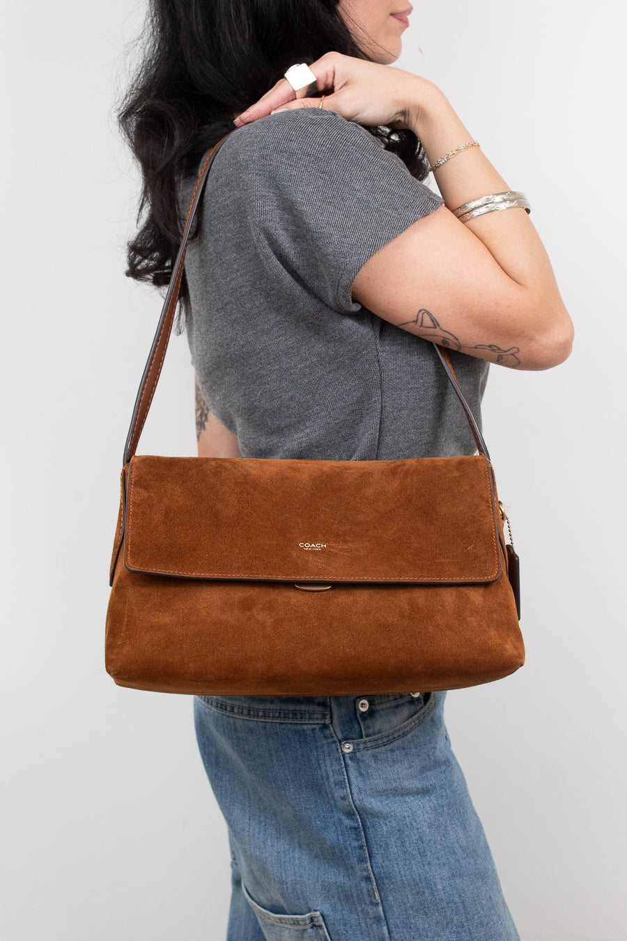 A woman wearing a gray t-shirt and blue jeans carries a brown suede shoulder bag with a flap closure and a small gold logo on the front, set against a plain light gray background.