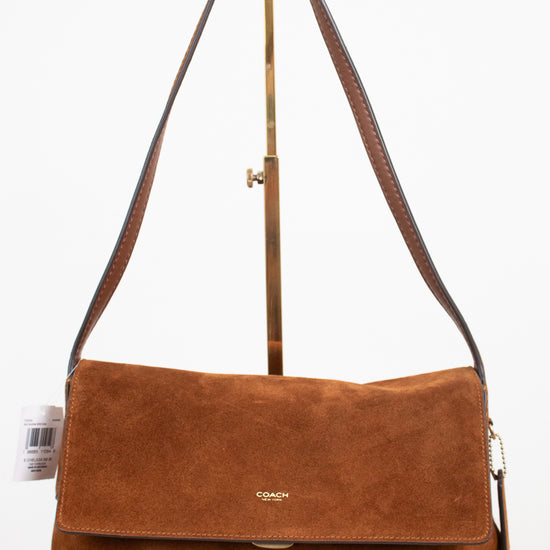 A brown suede Coach shoulder bag with a flap closure and a long shoulder strap, displayed on a stand against a white background.