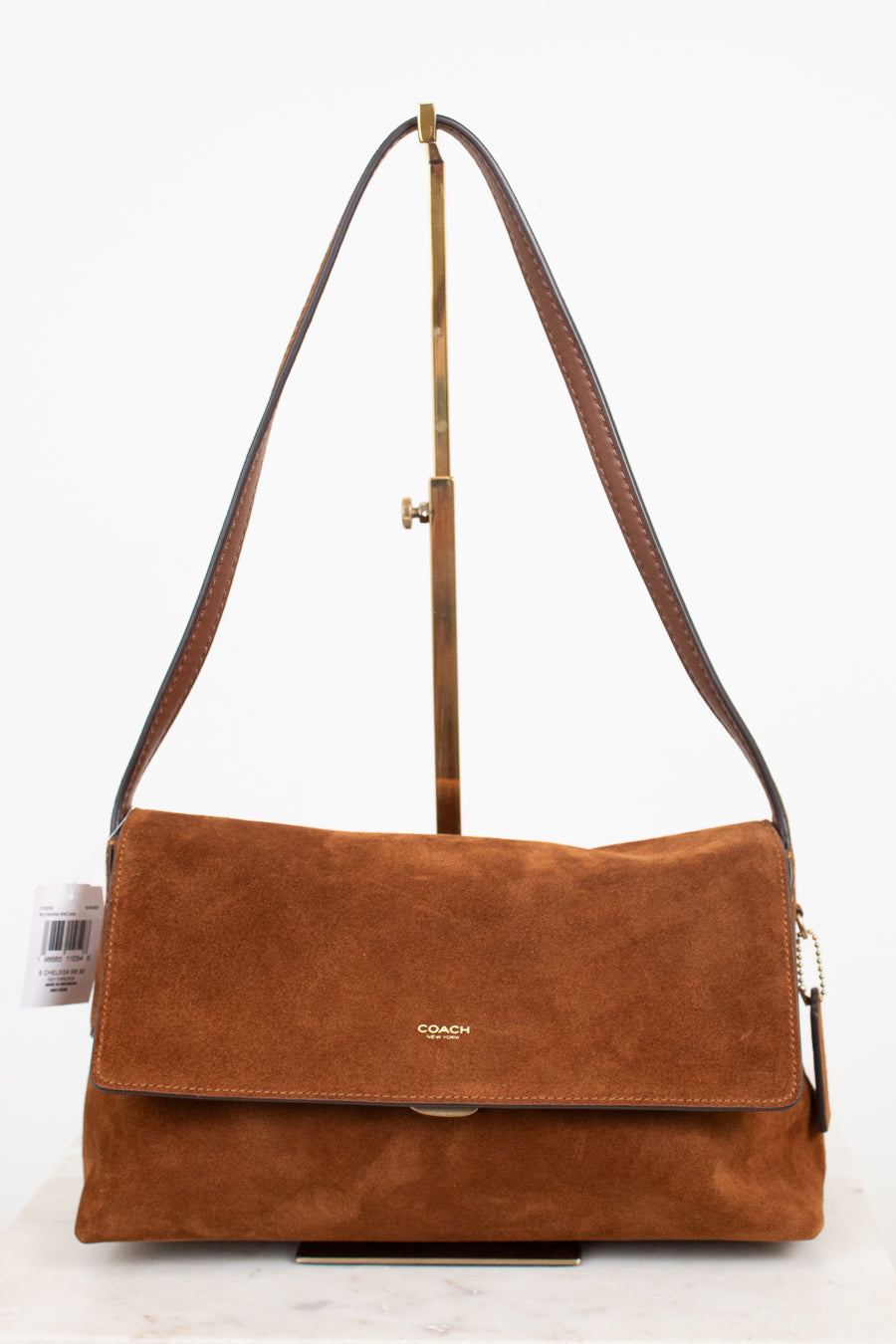 A brown suede Coach shoulder bag with a flap closure and a long shoulder strap, displayed on a stand against a white background.