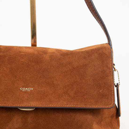 A Coach suede shoulder bag in a warm cognac brown, featuring a front flap with the gold 'COACH' logo and a dark brown leather strap, displayed on a minimalist stand against a white background.