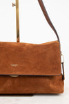 A Coach suede shoulder bag in a warm cognac brown, featuring a front flap with the gold 'COACH' logo and a dark brown leather strap, displayed on a minimalist stand against a white background.