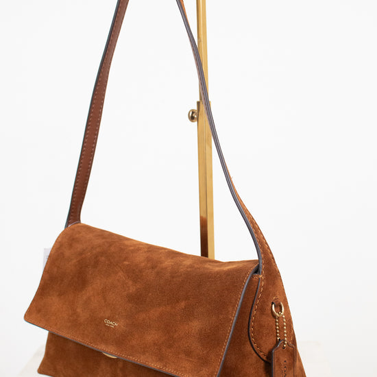 A brown suede shoulder bag with a flap closure and long strap, displayed on a gold metal stand against a white background.