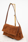 A brown suede shoulder bag with a flap closure and long strap, displayed on a gold metal stand against a white background.