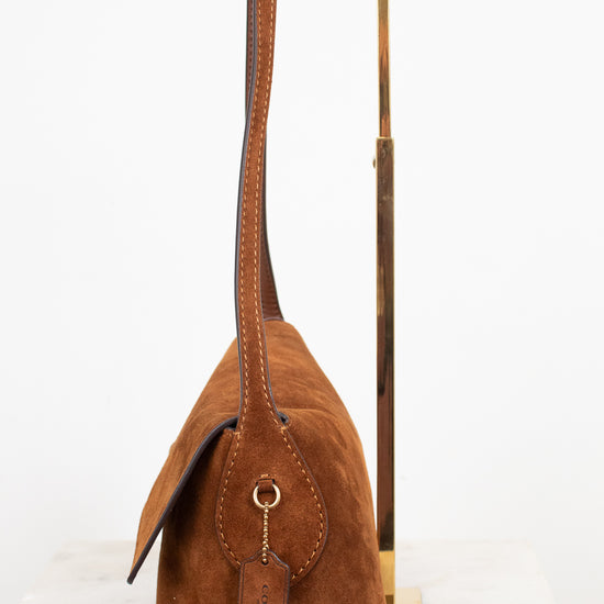 A brown suede shoulder bag with a long strap and a hanging leather tag, displayed on a minimalist gold metal stand against a white background.