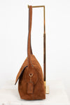 A brown suede shoulder bag with a long strap and a hanging leather tag, displayed on a minimalist gold metal stand against a white background.