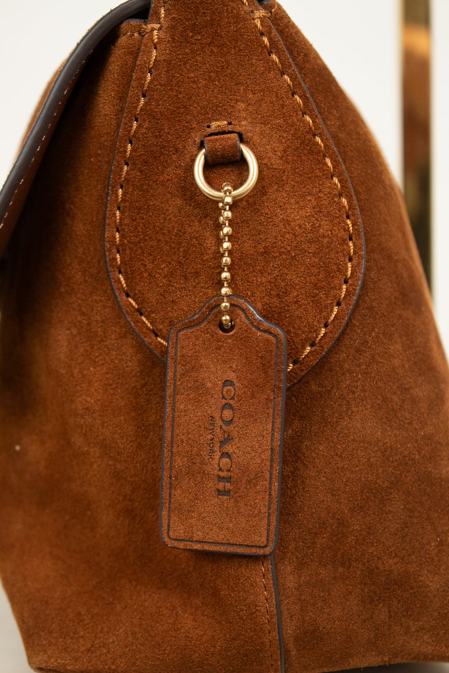 Close-up of a brown suede handbag with a gold-tone ring and chain holding a brown leather Coach hangtag.