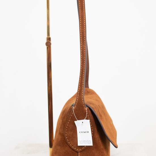 A camel brown suede shoulder bag with a long strap, displayed on a gold metal stand against a white background. A white paper tag with the brand name 'COACH' is attached.