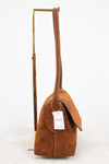 A camel brown suede shoulder bag with a long strap, displayed on a gold metal stand against a white background. A white paper tag with the brand name 'COACH' is attached.