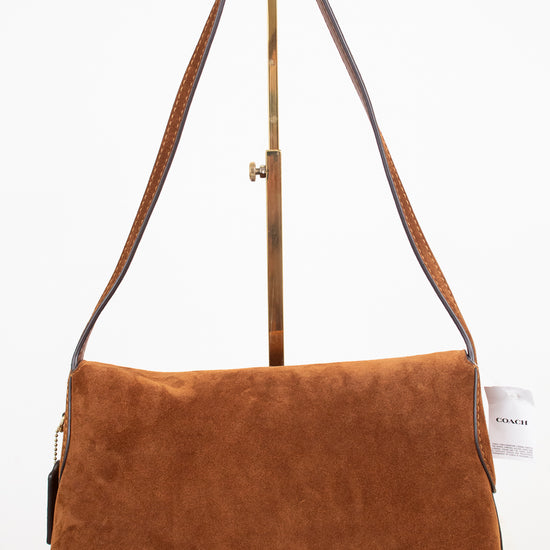 A brown suede shoulder bag with a long brown leather strap and a gold-tone metal stand, photographed against a clean white background.