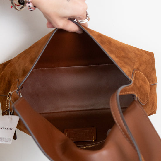 A close-up of a brown suede and leather Coach shoulder bag being held open by two hands, showing the interior lining and a Coach brand tag hanging from the zipper.