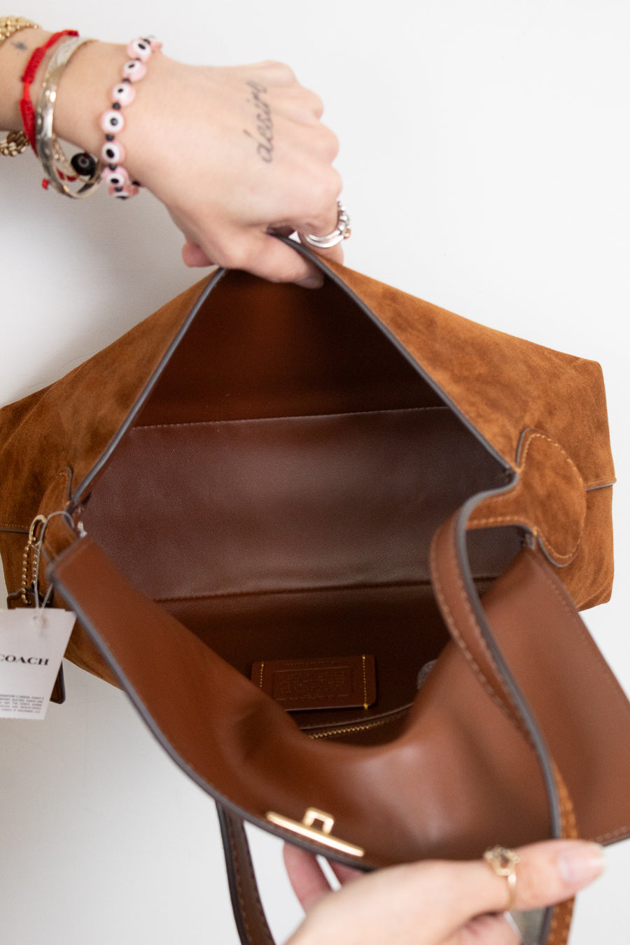 A close-up of a brown suede and leather Coach shoulder bag being held open by two hands, showing the interior lining and a Coach brand tag hanging from the zipper.
