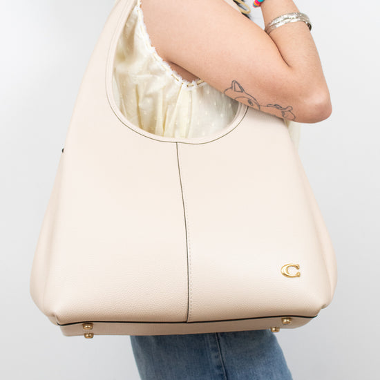A woman wearing a cream-colored leather hobo shoulder bag with gold-tone hardware, shown from the side against a light gray background.