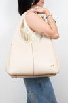 A woman wearing a cream-colored leather hobo shoulder bag with gold-tone hardware, shown from the side against a light gray background.