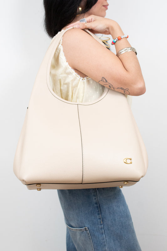 A woman wearing a cream-colored leather hobo shoulder bag with gold-tone hardware, shown from the side against a light gray background.