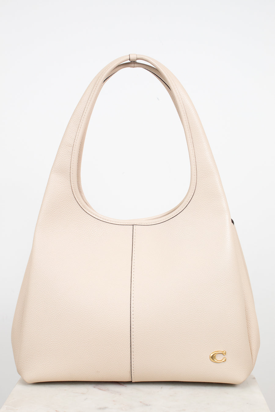 A light beige leather shoulder bag with, a vertical center seam, and a small gold logo emblem on the bottom right corner, set against a plain white background.
