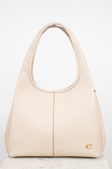 A light beige leather shoulder bag with, a vertical center seam, and a small gold logo emblem on the bottom right corner, set against a plain white background.