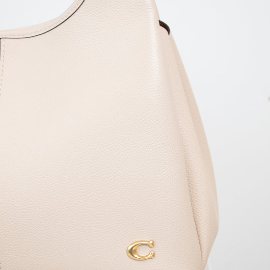 A close-up of a light beige pebbled leather handbag with a small gold-tone 'C' logo on the front, resting on a white marble surface against a white background.