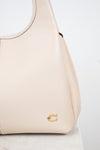 A close-up of a light beige pebbled leather handbag with a small gold-tone 'C' logo on the front, resting on a white marble surface against a white background.