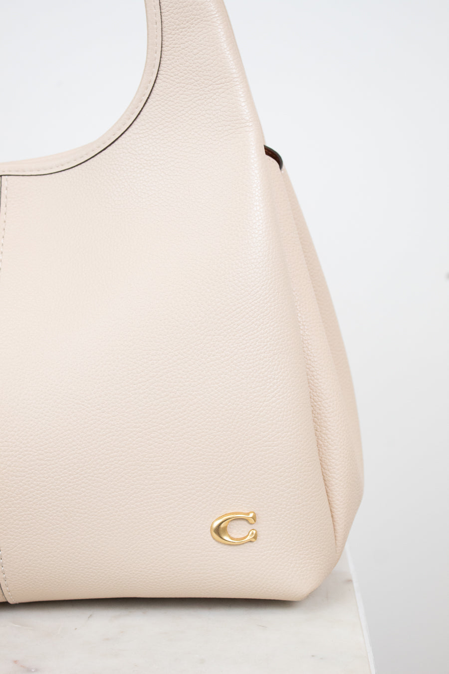 A close-up of a light beige pebbled leather handbag with a small gold-tone 'C' logo on the front, resting on a white marble surface against a white background.