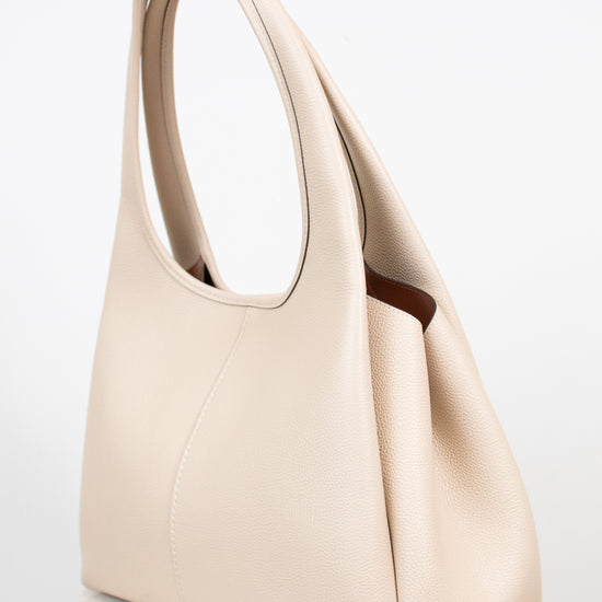 A light beige leather shoulder bag with a top handle and a small gold logo emblem near the bottom front, displayed on a light marble surface against a white background.