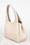 A light beige leather shoulder bag with a top handle and a small gold logo emblem near the bottom front, displayed on a light marble surface against a white background.