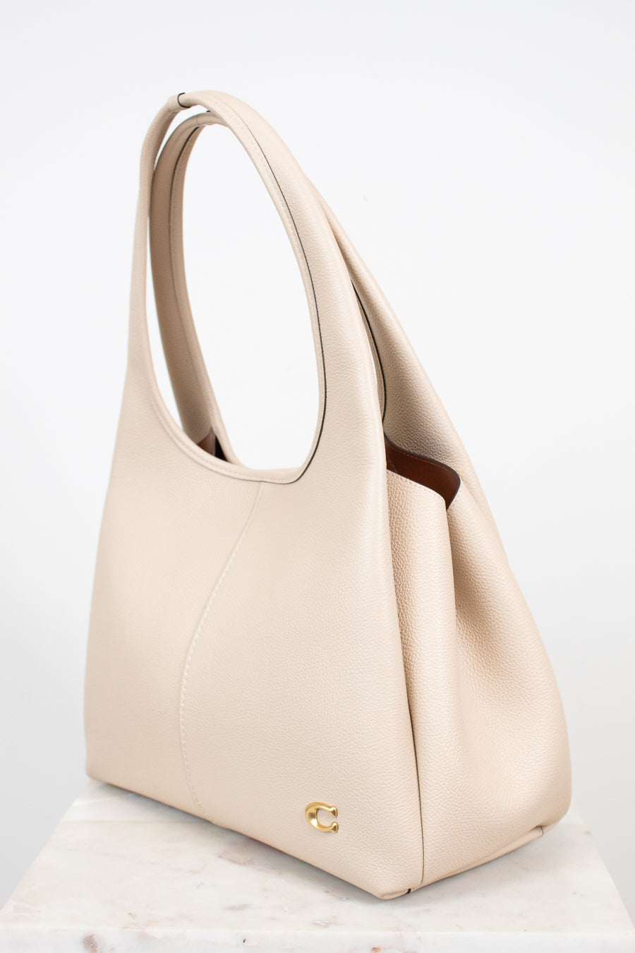 A light beige leather shoulder bag with a top handle and a small gold logo emblem near the bottom front, displayed on a light marble surface against a white background.