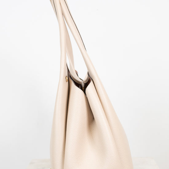 A light beige leather shoulder bag with long shoulder straps, standing on a white marble surface against a white background.