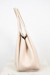 A light beige leather shoulder bag with long shoulder straps, standing on a white marble surface against a white background.