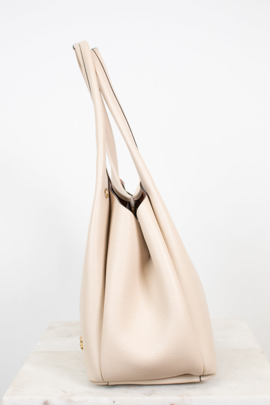 A light beige leather shoulder bag with long shoulder straps, standing on a white marble surface against a white background.