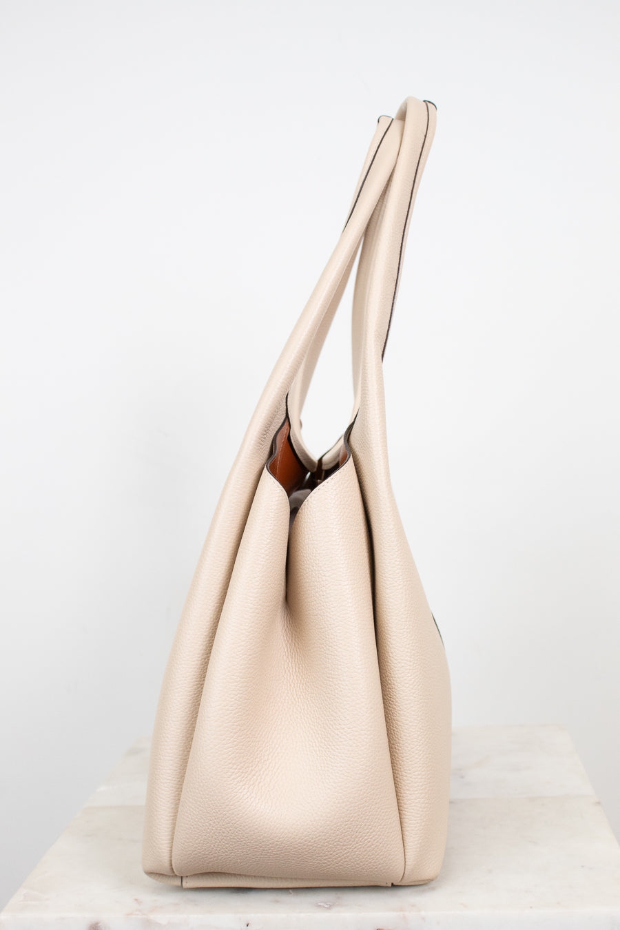 A light beige leather hobo handbag with long shoulder straps, standing upright on a light marble surface against a white background.