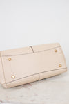 Bottom view of a beige leather shoulder bag with gold-tone metal feet, shown from a side angle on a light marble surface against a white background.