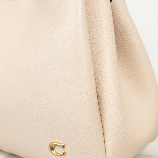 A close-up of a light beige pebbled leather handbag with a small gold-tone 'C' logo on the front, resting on a light marble surface.