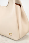 A close-up of a light beige pebbled leather handbag with a small gold-tone 'C' logo on the front, resting on a light marble surface.