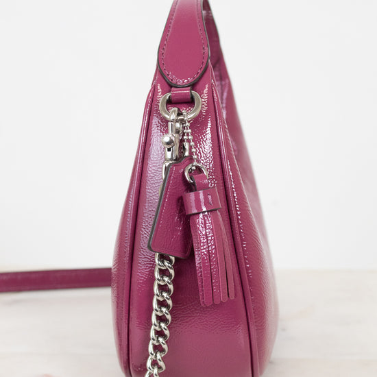A side profile view of a magenta pebbled leather shoulder bag with a silver chain strap and tassel detail, set against a plain white background.