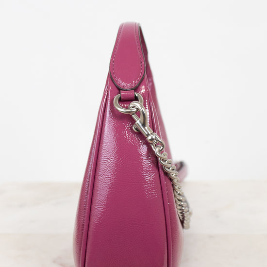 A close-up side view of a glossy magenta leather shoulder bag with a silver chain strap, resting on a light marble surface against a white background.