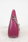 A close-up side view of a glossy magenta leather shoulder bag with a silver chain strap, resting on a light marble surface against a white background.