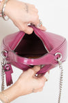 A close-up of a person's hands holding open a glossy magenta handbag with a silver chain strap and tassel zipper pull against a plain white background.