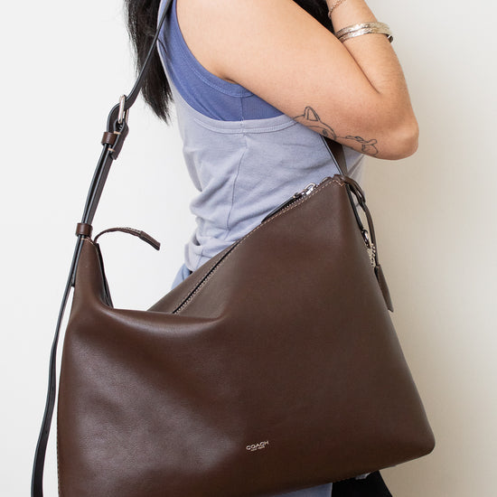 Model carrying a dark brown leather COACH shoulder bag with a structured, slouchy silhouette, a long adjustable strap , and a small silver logo on the front stating COACH.