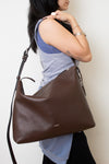 Model carrying a dark brown leather COACH shoulder bag with a structured, slouchy silhouette, a long adjustable strap , and a small silver logo on the front stating COACH.