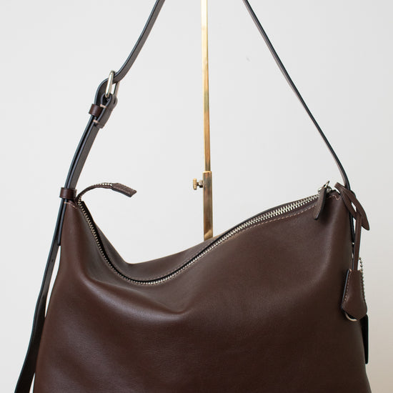 A dark brown leather hobo shoulder bag with a top zipper and a long shoulder strap, displayed on a white surface against a light gray background.