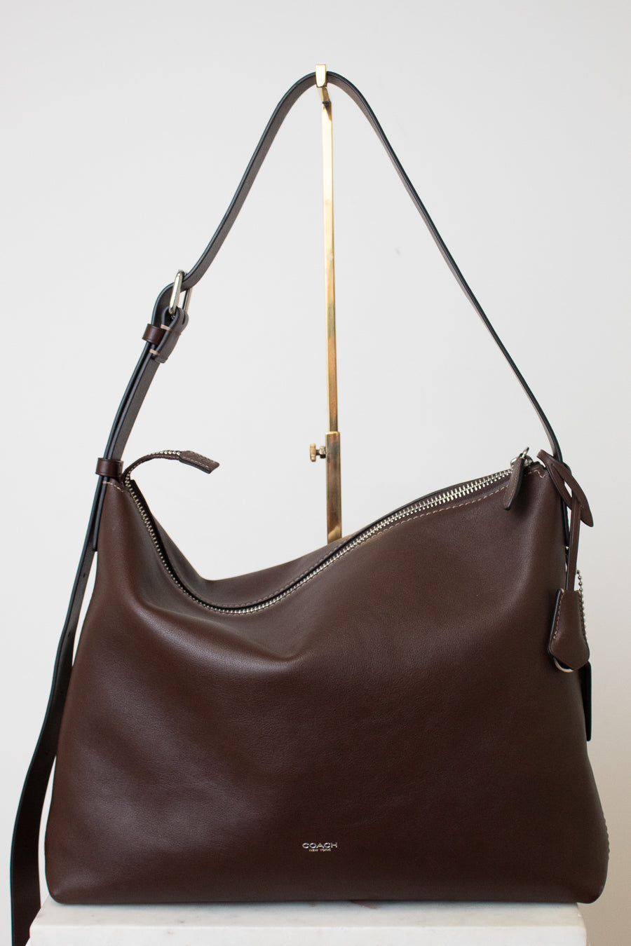 A dark brown leather hobo shoulder bag with a top zipper and a long shoulder strap, displayed on a white surface against a light gray background.