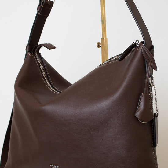 A dark brown leather shoulder bag with a slouchy hobo shape, displayed on a gold stand against a white background.