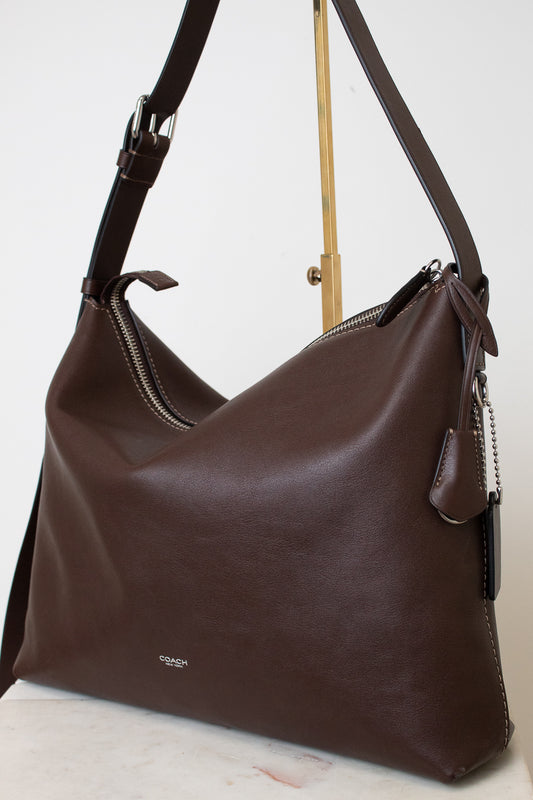 A dark brown leather shoulder bag with a slouchy hobo shape, displayed on a gold stand against a white background.