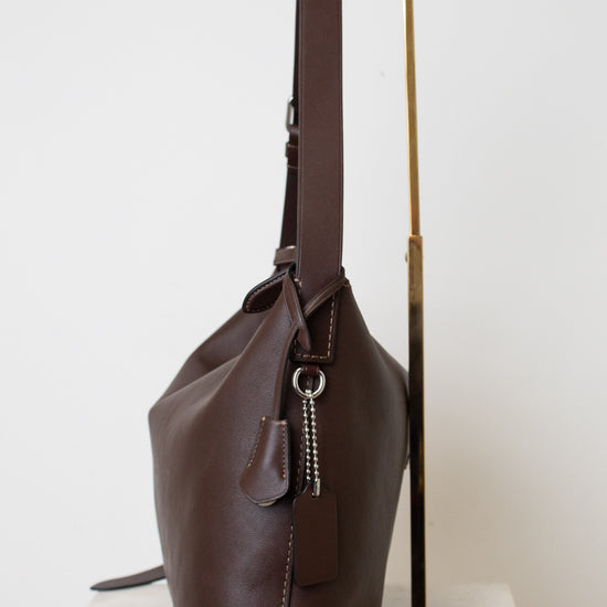 A dark brown leather shoulder bag with a long strap, displayed on a minimalist gold metal stand against a clean white background.