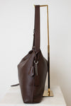 A dark brown leather shoulder bag with a long strap, displayed on a minimalist gold metal stand against a clean white background.