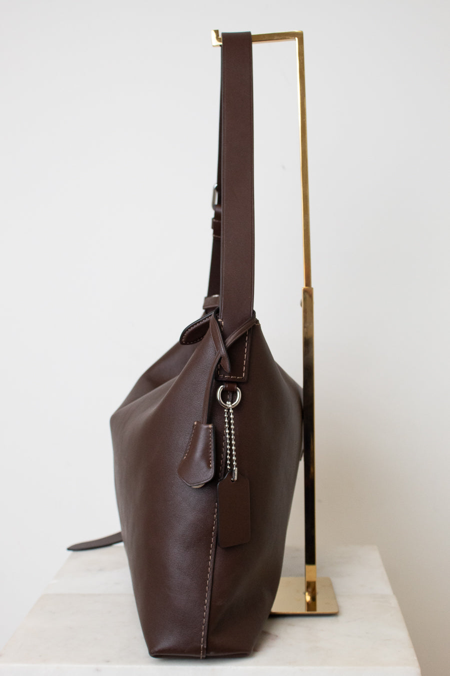 A dark brown leather shoulder bag with a long strap, displayed on a minimalist gold metal stand against a clean white background.