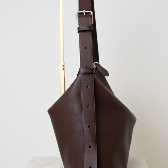 A dark brown leather bucket bag with a wide adjustable shoulder strap, displayed on a minimalist gold metal stand against a light gray background.
