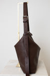 A dark brown leather bucket bag with a wide adjustable shoulder strap, displayed on a minimalist gold metal stand against a light gray background.